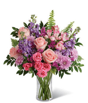 Floral arrangement featuring light pink roses, pink spray roses, pink alstroemeria, lavender stock, lavender mums, raspberry statice, and greenery in a glass vase.