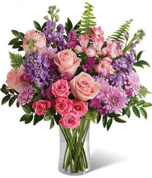 Illinois Floral arrangement featuring light pink roses, pink spray roses, pink alstroemeria, lavender stock, lavender mums, raspberry statice, and greenery in a glass vase.