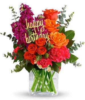 Arizona Floral arrangement featuring orange roses, orange spray roses, fuchsia stock, pink carnations, spiral eucalyptus, and lemon leaf with a 'Happy Birthday' pick in a clear cube vase.
