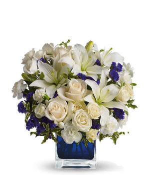 Floral arrangement featuring crme roses, crme spray roses, white Asiatic lilies, white miniature carnations, and purple statice in a blue cube vase.