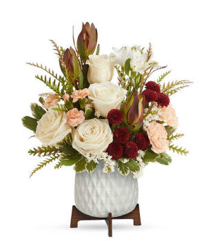A bouquet of cr&egrave;me roses, white alstroemeria, red leucadendron, peach carnations, and burgundy button spray chrysanthemums in a white vase with a wooden stand.